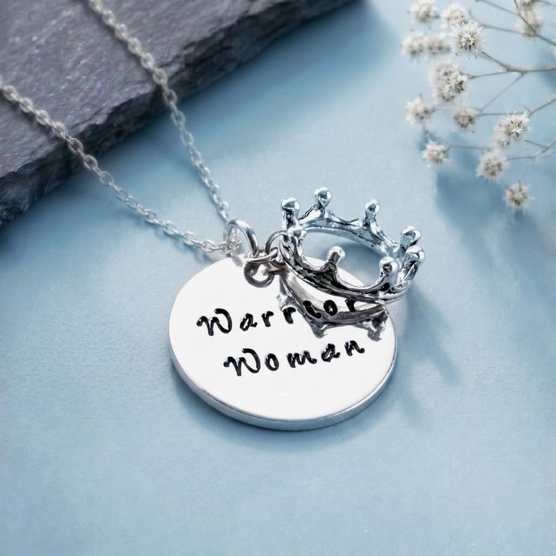 May include: A silver-toned necklace featuring a circular pendant engraved with the text Warrior Woman in black script. A small, detailed silver-toned crown charm is attached to the jump ring, resting above the circular pendant on a delicate chain.