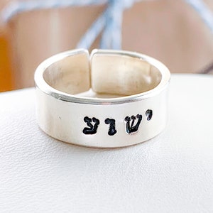 May include: A silver-colored open-band ring featuring a smooth, polished finish. The outer surface of the band is engraved with three distinct black Hebrew characters, creating a clear contrast against the metallic silver background of the ring.
