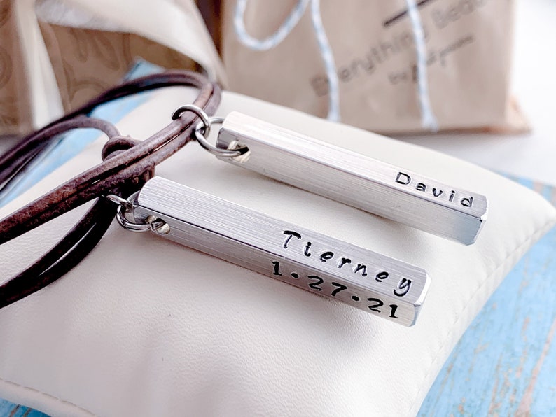 A silver 3D rectangle-shaped pendant with four sides sits on a jewelry display pillow. The front side of the pendant is hand stamped with a personalized name. It hangs from a brown or black leather cord with sliding knots for adjusting.