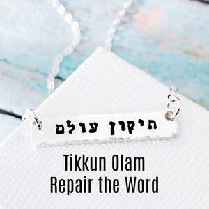 May include: Silver necklace with a bar pendant that reads "Tikkun Olam" in Hebrew. The pendant is inscribed with the Hebrew words "Tikkun Olam" which translates to "Repair the World".
