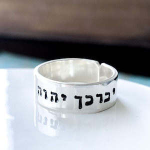 May include: A silver-colored band ring with an open back design. The outer surface of the ring features black engraved Hebrew text that reads יברכך יהוה. The metal has a smooth, polished finish, and the ring is shown from a slightly elevated angle.