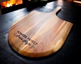 Könnte beinhalten: Ein Pizzaschieber aus Holz mit einer abgerundeten Klinge und einem Griff. Die Oberfläche weist eine natürliche Holzmaserung auf und ist mit dem Text "POWERS FAMILY Pizza Night" sowie zwei kleinen Pizzastück-Symbolen lasergraviert.