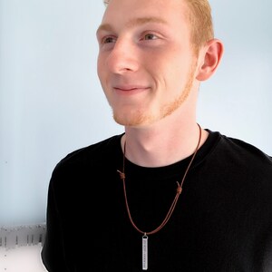 The handsome, red headed male model is smiling and wearing a black shirt with a necklace dangling on his chest.  The necklace is a brown cord with sliding knots and has a 3D, rectangle pendant. The pendant is long and about 1 3/4 inches.