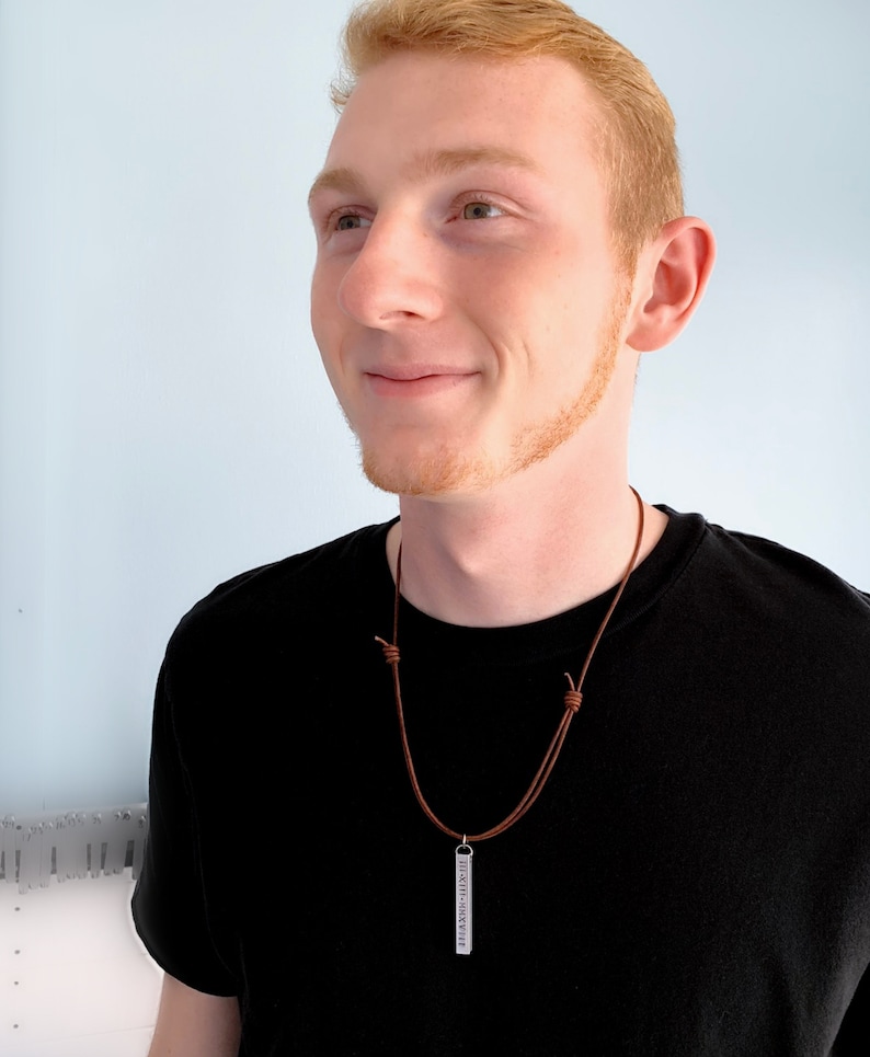 The handsome, red headed male model is smiling and wearing a black shirt with a necklace dangling on his chest.  The necklace is a brown cord with sliding knots and has a 3D, rectangle pendant. The pendant is long and about 1 3/4 inches.