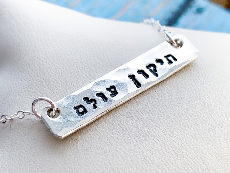May include: Silver necklace with a hammered bar pendant featuring the Hebrew words "עוצם" and "חזק" in black lettering.