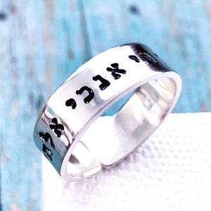 May include: A silver-toned metal ring featuring a wide band with an open back design. The outer surface of the band is engraved with black Hebrew characters that wrap around the circumference of the ring, creating a distinct contrast against the metal.