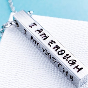 A silver, 3D, rectangle pendant on a display box. The front side of the pendant is hand stamped with the words I am enough in a cute font. The other sides read I am strong, I am loved, and I am worthy. It hangs from a silver regular chain.