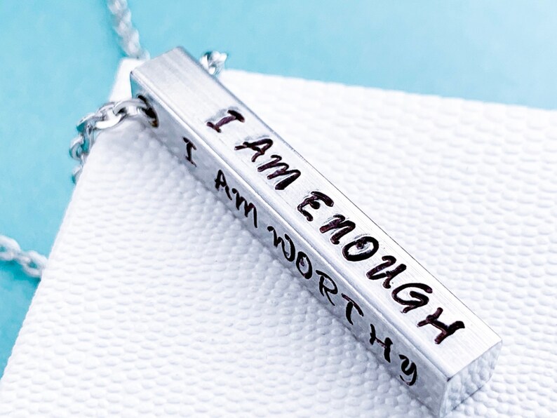 A silver, 3D, rectangle pendant on a display box. The front side of the pendant is hand stamped with the words I am enough in a cute font. The other sides read I am strong, I am loved, and I am worthy. It hangs from a silver regular chain.