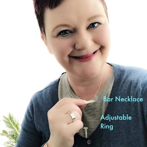 May include: A woman is wearing a blue sweater, a silver bar necklace with the word "life" engraved on it, a silver ring, and a silver 1/4 inch cuff bracelet. The necklace is labeled "Bar Necklace", the ring is labeled "Adjustable Ring", and the bracelet is labeled "1/4" Cuff Bracelet".
