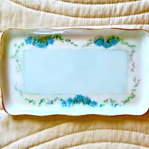 May include: A rectangular porcelain tray with a scalloped edge and a gold-painted rim. The center features a light blue rectangular panel bordered by white dots, surrounded by hand-painted blue forget-me-not flowers and green foliage.
