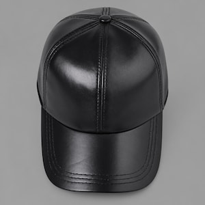 Puede incluir: Una gorra de béisbol negra confeccionada con un material liso y semibrillante. La gorra presenta un diseño de seis paneles con costuras visibles que irradian desde un botón central en la corona y una visera curva con tres filas de costuras.