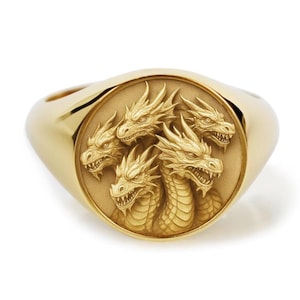 May include: A gold signet ring featuring a circular face with a detailed, raised relief carving of a multi-headed dragon. The dragon heads are arranged in a cluster, showing scales, sharp teeth, and horns, all rendered in a polished gold finish.