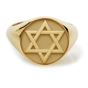 May include: A gold signet ring featuring a circular face with a raised Star of David symbol. The center of the ring has a matte, textured finish, while the outer band and the edges of the star have a polished, reflective gold surface.