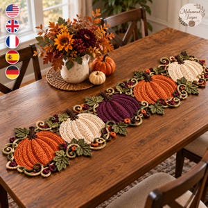 Puede incluir: Un camino de mesa de ganchillo rectangular con un patrón repetitivo de calabazas en naranja, crema y morado oscuro, acentuado con hojas verdes y enredaderas marrones tejidas, colocado sobre una mesa de madera oscura.