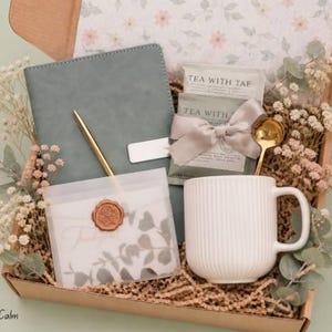 May include: A gift box containing a sage green notebook, a gold pen, a white ribbed ceramic mug, a gold spoon, and tea packets labeled Tea With Tae. A translucent envelope with a wax seal and dried flowers are included in the cardboard box.