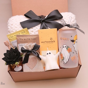 May include: A gift box containing a white knit blanket with a black bow, two Tea With Tae packets, a Moonstruck chocolate bar, a glass tumbler with ghost graphics, a honey dipper, a black rose, a burlap pouch, and a ghost-shaped candle.