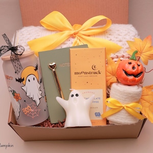 May include: A gift box containing a frosted glass tumbler with ghost graphics, a green notebook, a gold pen with a heart topper, a Moonstruck chocolate bar, a white ghost candle, a small pumpkin, a white knit item, and yellow ribbon accents.