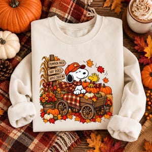May include: A cream-colored crewneck sweatshirt featuring a graphic of Snoopy wearing an orange cap and scarf, sitting in a wooden wagon filled with pumpkins. A signpost reads PUMPKIN PATCH, HAY RIDES, and APPLE CIDER, surrounded by autumn leaves.