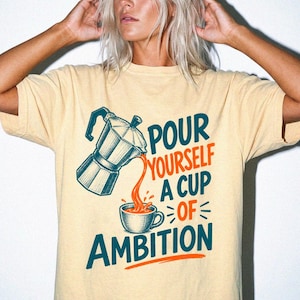 May include: A light yellow short-sleeve t-shirt featuring a graphic of a moka pot pouring liquid into a cup. The text reads "POUR YOURSELF A CUP OF AMBITION" in dark teal and orange lettering. The design is centered on the front of the garment.