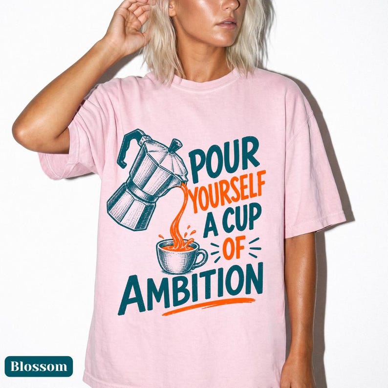 May include: A light pink t-shirt featuring a graphic of a moka pot pouring liquid into a coffee cup. The text reads, "POUR YOURSELF A CUP OF AMBITION." The design uses dark teal and orange ink, with the moka pot and cup rendered in a detailed, etched style.