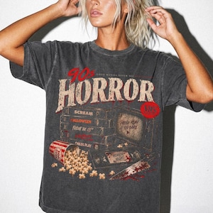 Op de afbeelding: Een antracietgrijs t-shirt met een grafische print met de tekst 90s HORROR, GOOD MOVIES NEVER DIE en PRESS PLAY IF YOU DARE. Het ontwerp bevat een stapel VHS-banden, een emmer popcorn, een mes en een televisiescherm.