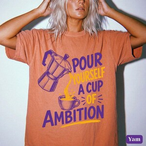 May include: A terracotta-colored short-sleeve t-shirt featuring a graphic of a moka pot pouring coffee into a cup. The text reads "POUR YOURSELF A CUP OF AMBITION" in purple and yellow lettering. The design is centered on the front of the shirt.