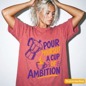 May include: A watermelon-colored short-sleeve t-shirt featuring a graphic of a moka pot pouring coffee into a cup. The text reads, "POUR YOURSELF A CUP OF AMBITION" in purple and yellow lettering. The design is centered on the front of the shirt.