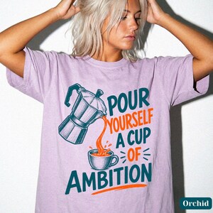May include: A light purple short-sleeve t-shirt featuring a graphic of a moka pot pouring coffee into a cup. The text reads "POUR YOURSELF A CUP OF AMBITION" in dark teal and orange lettering. The design is centered on the front of the garment.