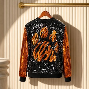 May include: A long-sleeve jacket covered in black and orange sequins. The back panel features a large, abstract tiger paw print design created with orange sequins against a black sequined background. The sleeves are solid orange sequins.