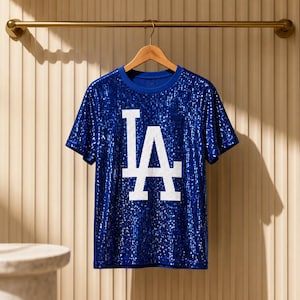 May include: A short-sleeved t-shirt covered in royal blue sequins featuring a large white sequined LA logo on the front. The garment is displayed on a wooden hanger suspended from a horizontal metal rod against a vertical paneled wall.