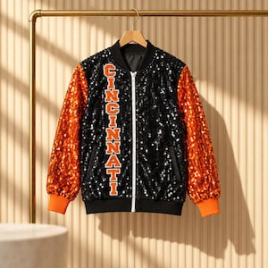 Könnte beinhalten: Eine schwarze Pailletten-Bomberjacke mit leuchtend orangefarbenen Paillettenärmeln und gerippten orangefarbenen Bündchen. Auf der Vorderseite steht vertikal das Wort CINCINNATI in orangefarbenen Blockbuchstaben mit weißem Rand. Mit weißem Reißverschluss.