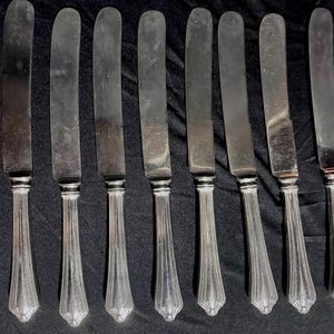 May include: A set of eight silver-toned metal dinner knives arranged in a row. Each knife features a smooth, rounded blade and a handle decorated with vertical fluted lines that taper toward the base, creating a uniform and classic appearance.