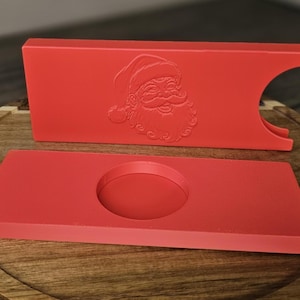 May include: A red rectangular stand with a Santa Claus face embossed on the front and a circular cutout on the side, paired with a matching red rectangular base featuring a centered circular indentation for holding items securely.