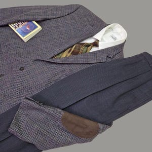 Può includere: Un blazer grigio con motivo pied-de-poule e toppe sui gomiti in camoscio marrone, una camicia bianca con colletto e una cravatta a quadri marroni, verdi e gialli. Un piccolo libro con copertina blu e il testo "РИХ" è appoggiato sul blazer. Pantaloni grigio scuro.