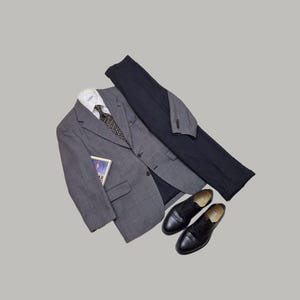 Puede incluir: Un blazer gris, una camisa blanca con cuello, una corbata oscura estampada, un pantalón azul marino oscuro y zapatos de vestir de cuero negro. Un pequeño libro rectangular con una cubierta azul y morada está metido en el bolsillo del blazer.