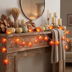 May include: A string of LED lights featuring orange and red maple leaf covers draped across a wooden mantel. The display includes several small pumpkins, a bundle of dried wheat, and pillar candles, creating a warm, illuminated autumn arrangement.