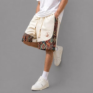 Puede incluir: Pantalones cortos de color blanco roto con cintura de cordón con borlas beige y un patrón geométrico colorido a lo largo de los paneles laterales y el dobladillo. Un pequeño parche cuadrado con un diseño gráfico es visible en la pierna izquierda, junto con zapatillas blancas.