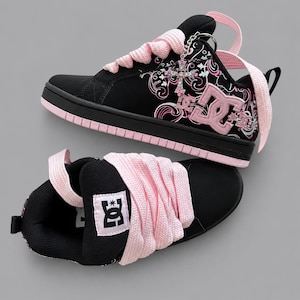 Puede incluir: Un par de zapatillas DC negras y rosa claro con cordones anchos y gruesos de color rosa claro y un logotipo DC prominente en el lateral. El panel lateral muestra un patrón gráfico decorativo de remolinos blancos y rosas y un dije de cruz plateada.