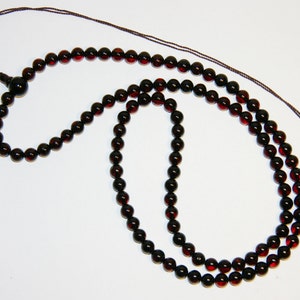 Puede incluir: Un collar de cuentas de color rojo oscuro con un cordón marrón. El collar está hecho de cuentas redondas y tiene un borla al final.