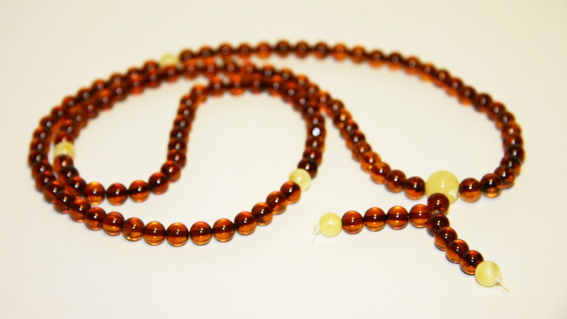 Pode incluir: Um colar de contas de mala de &acirc;mbar marrom com um tassel e duas contas brancas. O colar &eacute; feito de contas redondas e &eacute; enfiado em um cord&atilde;o.