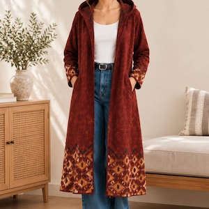 May include: A long, hooded, dark red coat featuring a subtle tonal pattern and a wide, geometric, multi-colored border at the hem and cuffs. The coat is worn over a white tank top, blue denim jeans with a black belt, and white sneakers.