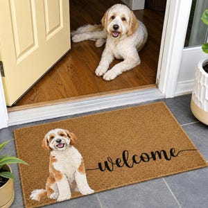 Puede incluir: Un felpudo rectangular de fibra de coco color canela con una ilustración impresa de un perro blanco y marrón sentado en el lado izquierdo. La palabra welcome está escrita en cursiva negra en el lado derecho de la alfombra, centrada.