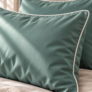 May include: A rectangular pillowcase in a solid muted teal green color featuring a crisp white piped edge trim along the perimeter. The fabric has a smooth, matte finish and is shown in a set of two, with one pillowcase positioned in the foreground.