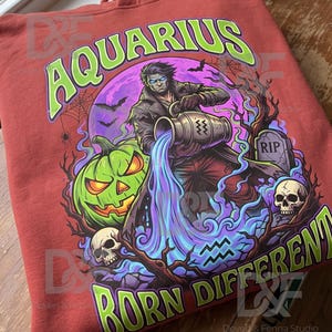 Op de afbeelding: Een roestkleurige hoodie met een grafische print van een figuur die water uit een kruik giet, omringd door een groene pompoen, schedels, een grafsteen en vleermuizen. De tekst luidt bovenaan AQUARIUS en onderaan BORN DIFFERENT.