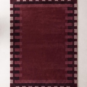 Op de afbeelding: Een rechthoekig vloerkleed met een effen diepbordeauxrood middenpaneel. De rand bestaat uit afwisselende donkerbordeauxrode en licht mauve rechthoekige blokken, wat een gestructureerd, herhalend geometrisch patroon rondom de gehele omtrek vormt.