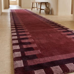 May include: A long rectangular runner rug featuring a deep burgundy center and a geometric border pattern in alternating shades of dark purple, plum, and light mauve. The rug has a dense, soft-textured pile and a clean, straight-edged finish.