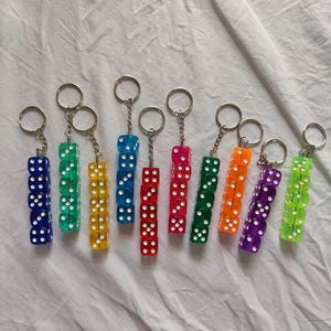 Puede incluir: Una colección de diez llaveros, cada uno con una pila vertical de cuatro dados translúcidos y coloridos con puntos blancos. Los colores de los dados incluyen azul, verde, amarillo, rojo, rosa, naranja, morado y verde lima, unidos a anillas de metal plateado.