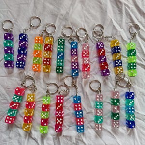 Pode incluir: Uma coleção de porta-chaves, cada um com uma pilha vertical de quatro dados de plástico translúcido e multicolorido com pontos brancos. Cada porta-chaves está preso a um anel metálico circular, exibindo várias combinações de cores, incluindo roxo, laranja, amarelo, verde, azul, rosa e vermelho.