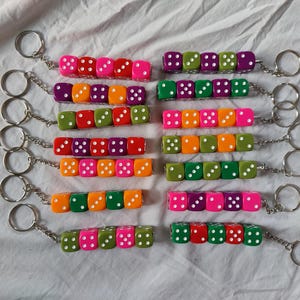 Può includere: Una collezione di quattordici portachiavi, ognuno composto da una fila di cinque dadi in plastica colorata fissati a un anello in metallo argentato tramite una corta catenella. I dadi presentano varie combinazioni di colori tra cui rosa, arancione, verde, viola e rosso.