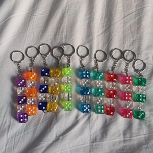 Pode incluir: Uma coleção de onze porta-chaves, cada um com uma pilha vertical de três dados translúcidos coloridos com pontos brancos. Os dados estão ligados por uma corrente de metal prateado e um anel circular, apresentando várias cores, incluindo roxo, laranja, azul, amarelo, verde-limão, turquesa, vermelho, rosa e verde-escuro.